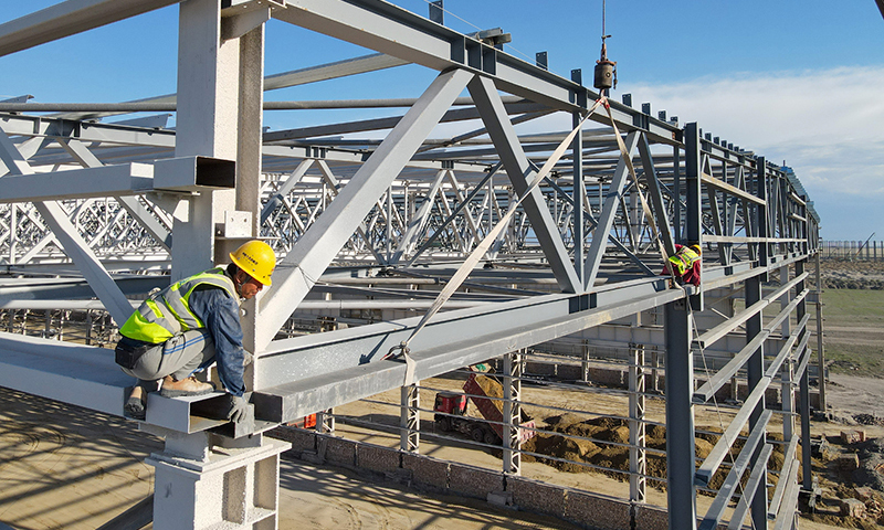 進入施工旺季，新疆塔城重點開發開放試驗區工程建設有序進行，來自中鐵十五局等多家單位的工人晝夜奮戰在建設現場。新疆塔城重點開發開放試驗區是我國西北地區第一個沿邊重點開發開放試驗區。圖為工人在20余米高的新疆塔城重點開發開放試驗區一處風機葉片制造項目頂部進行屋面檁條安裝作業（5月18日無人機拍攝）。新華社記者 高晗 攝