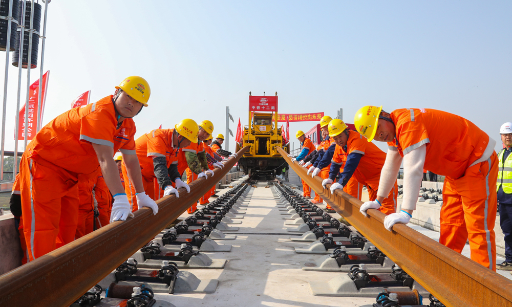 為優質高效完成鋪軌任務，建設者提前在位于荊州站北側的鋪軌基地儲存長鋼軌，以鋪軌基地為起點，采用TC-500型鋪軌機由荊州站一路向北鋪設長鋼軌；加持智能行車調度指揮系統，全天候對鋪軌機、長軌運輸車、焊軌車等設備的運行狀態及作業位置進行監控和統一調度指揮，保證了交叉施工安全有序，并且提高了鋪軌速度，每天單向鋪軌可達6公里。圖為鋪軌作業現場，作業人員在鋪軌機的牽引下正在安裝軌道。