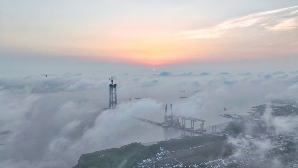 清晨，建設中的江蘇張靖皋長江大橋雨后初霽，旭日照亮云層，大橋雛形若隱若現，美輪美奐。目前，中國交建旗下中交二航局承建的張靖皋長江大橋南主塔建設高度已達220多米，預計2025年底可實現封頂，屆時高度將達350米，為世界最高懸索橋索塔。